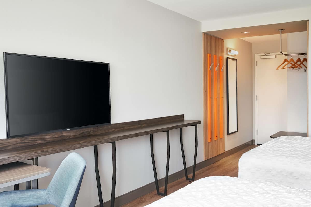 A hotel room with a flat-screen TV, a wooden desk, a light blue chair, a bed with white bedding, and a wooden wardrobe with a mirror.