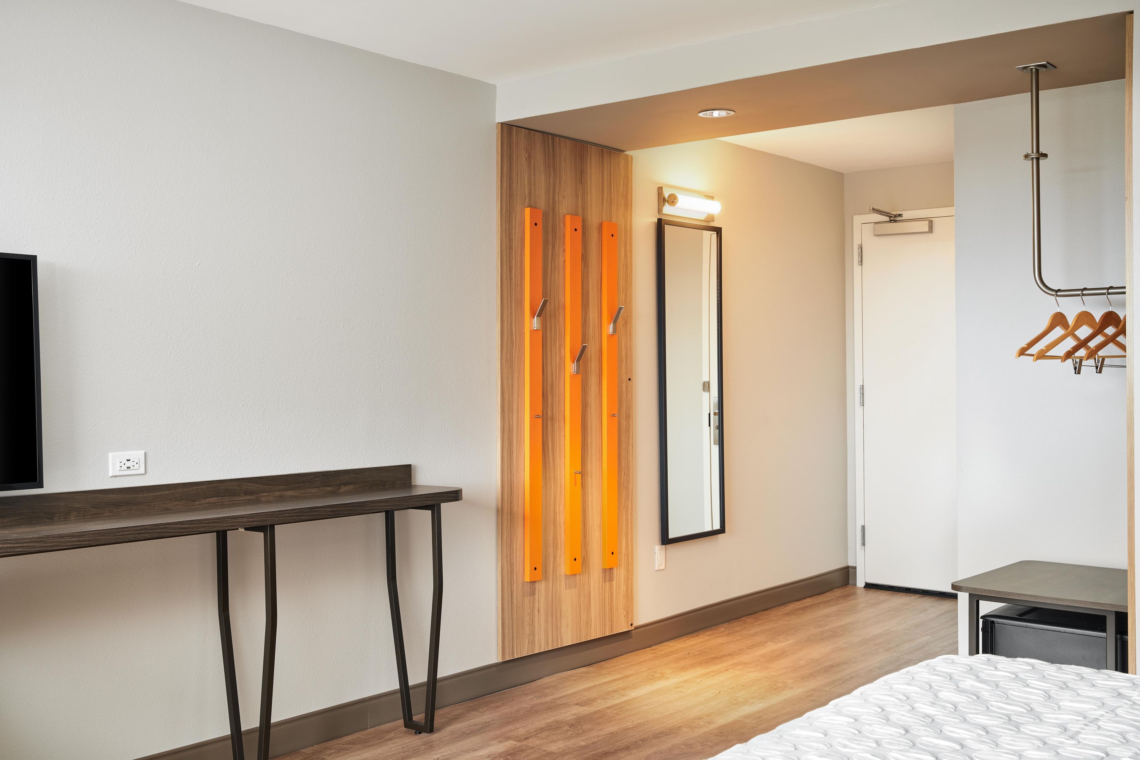 A modern hotel room with a wooden wardrobe, a mirror, a coat rack, and a bed with a textured mattress.