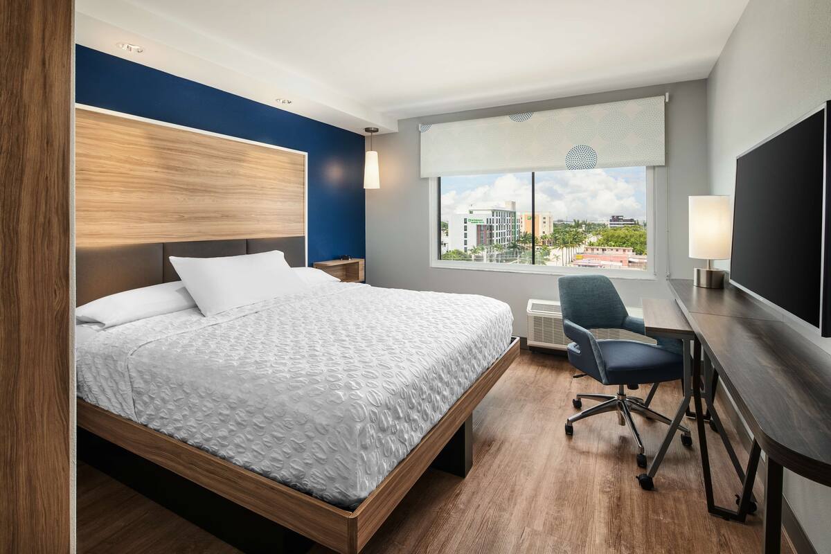 A modern hotel room with a large bed, a desk, and a chair. There is a window with a view of buildings and a cityscape.