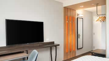 A hotel room with a flat-screen TV, a desk, a chair, a bed, a wardrobe, a mirror, and a coat rack.