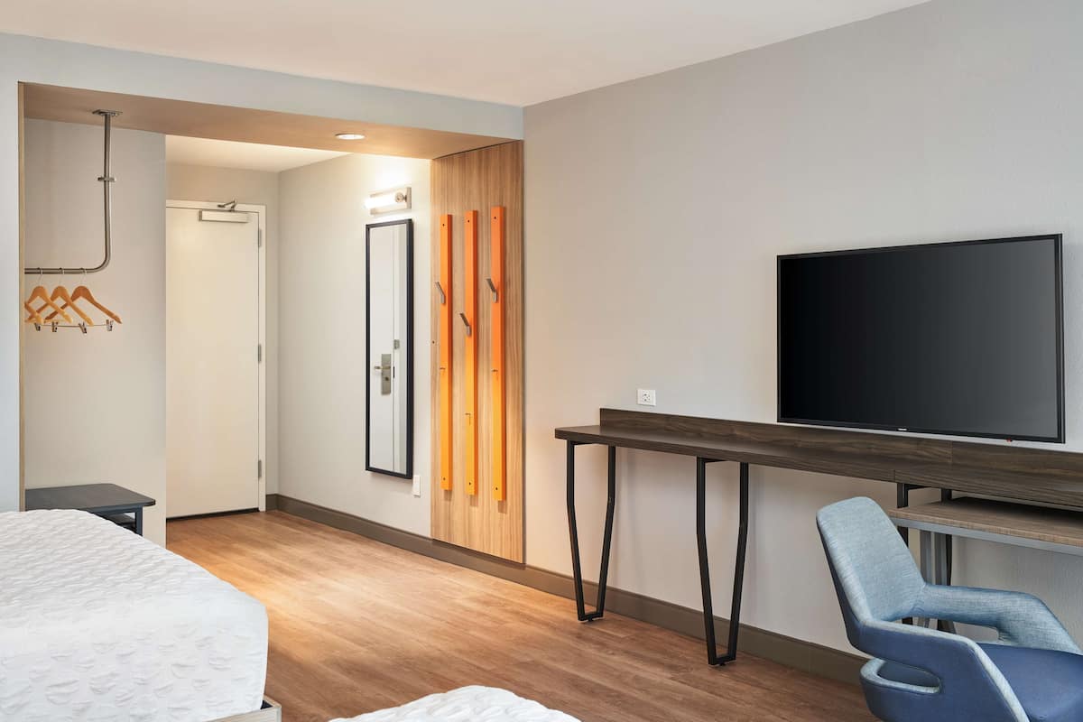 A modern hotel room with a bed, a desk with a flat-screen TV, a chair, and a wooden wardrobe.