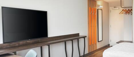 A hotel room with a flat-screen TV, a wooden desk, a chair, a bed, a wardrobe, and a mirror.
