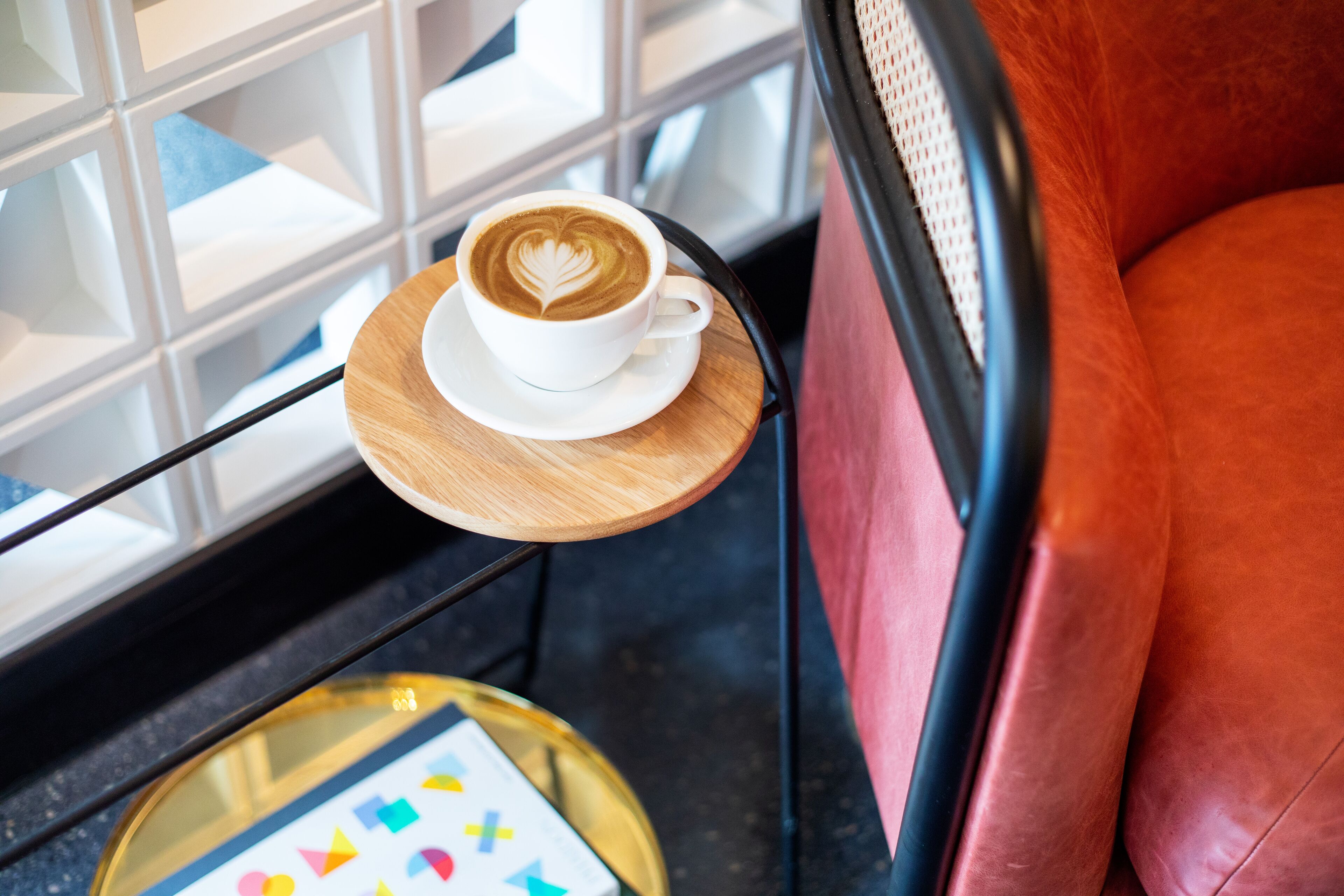 A cup of coffee with a heart design on a saucer, placed on a wooden table next to a red leather couch.