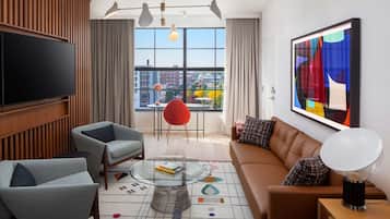 A modern living room with a large TV, comfortable seating, a glass coffee table, and a city view through large windows.