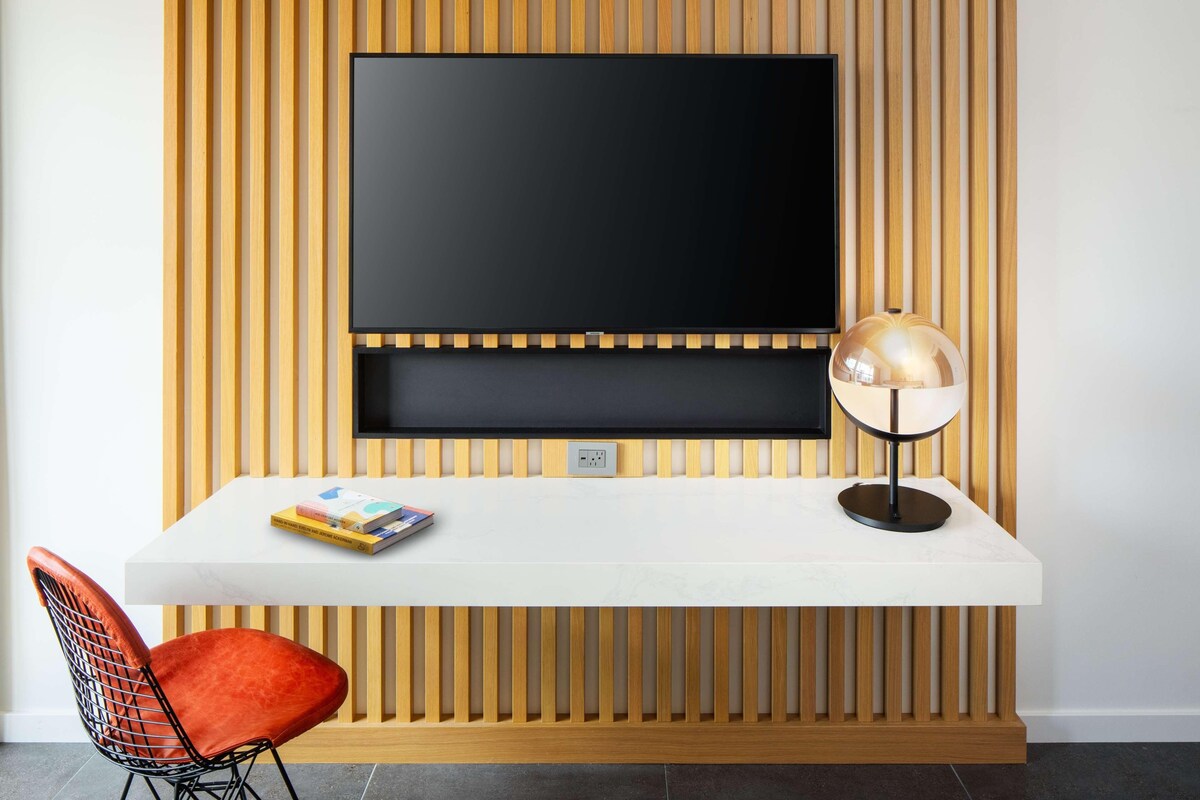 A modern living room with a flat-screen TV mounted on a wooden panel wall, a white console table with a lamp, and a red chair.