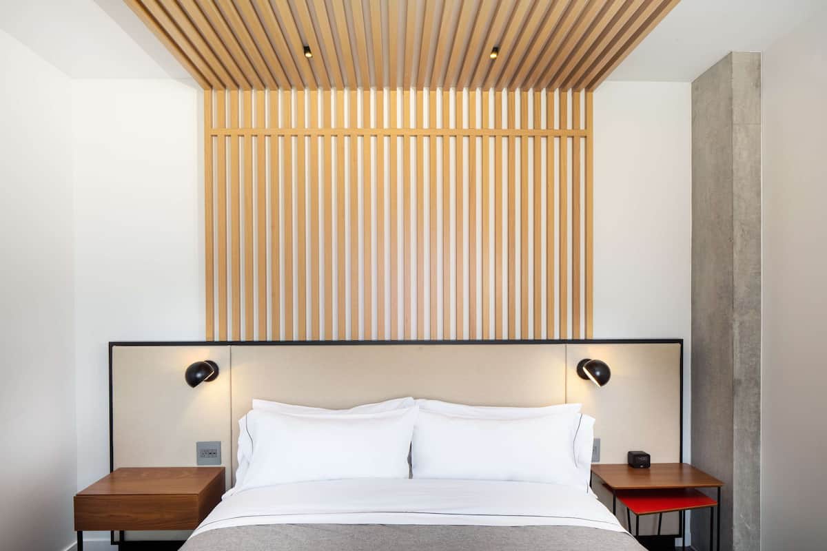 Una habitación de hotel moderna con una cama grande, cabecera de madera, dos mesitas de noche y una mesita auxiliar roja.