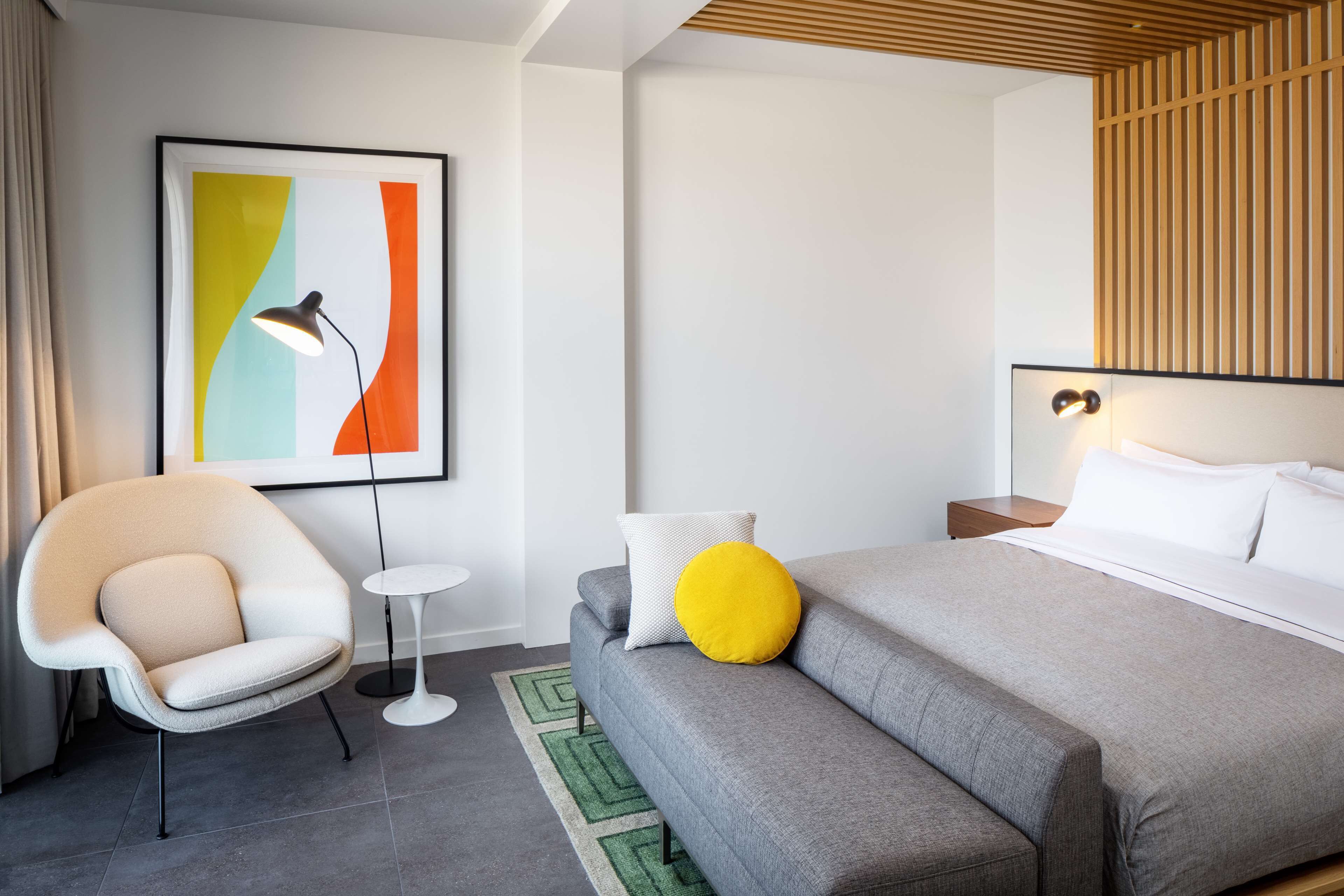 Un hotel moderno con un sofá gris, una mesita redonda pequeña, una silla, una cama con almohadas blancas y amarillas, y un cuadro abstracto colorido en la pared.
