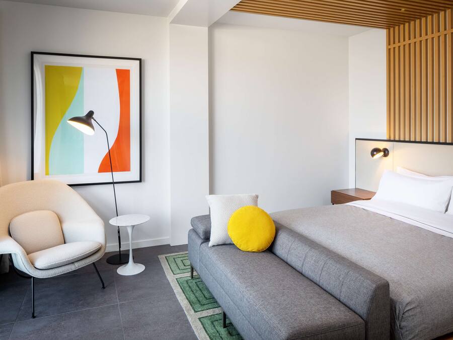 A modern hotel room with a grey sofa, a small round table, a chair, a bed with white and yellow pillows, and a colorful abstract painting on the wall.