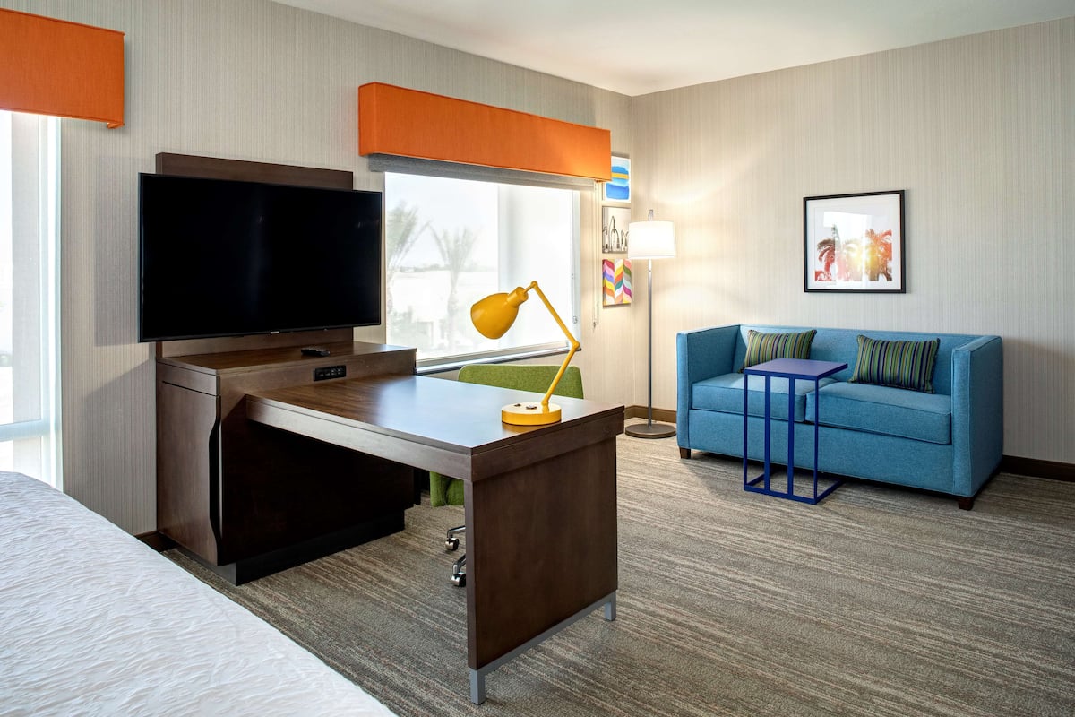 A hotel room with a bed, a desk with a TV, a desk lamp, a blue sofa, and a window with a view.