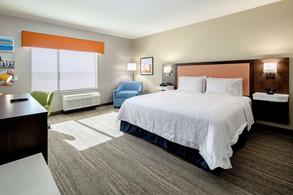 A hotel room with a large bed, a desk, a chair, a window with blinds, and a wall-mounted lamp.