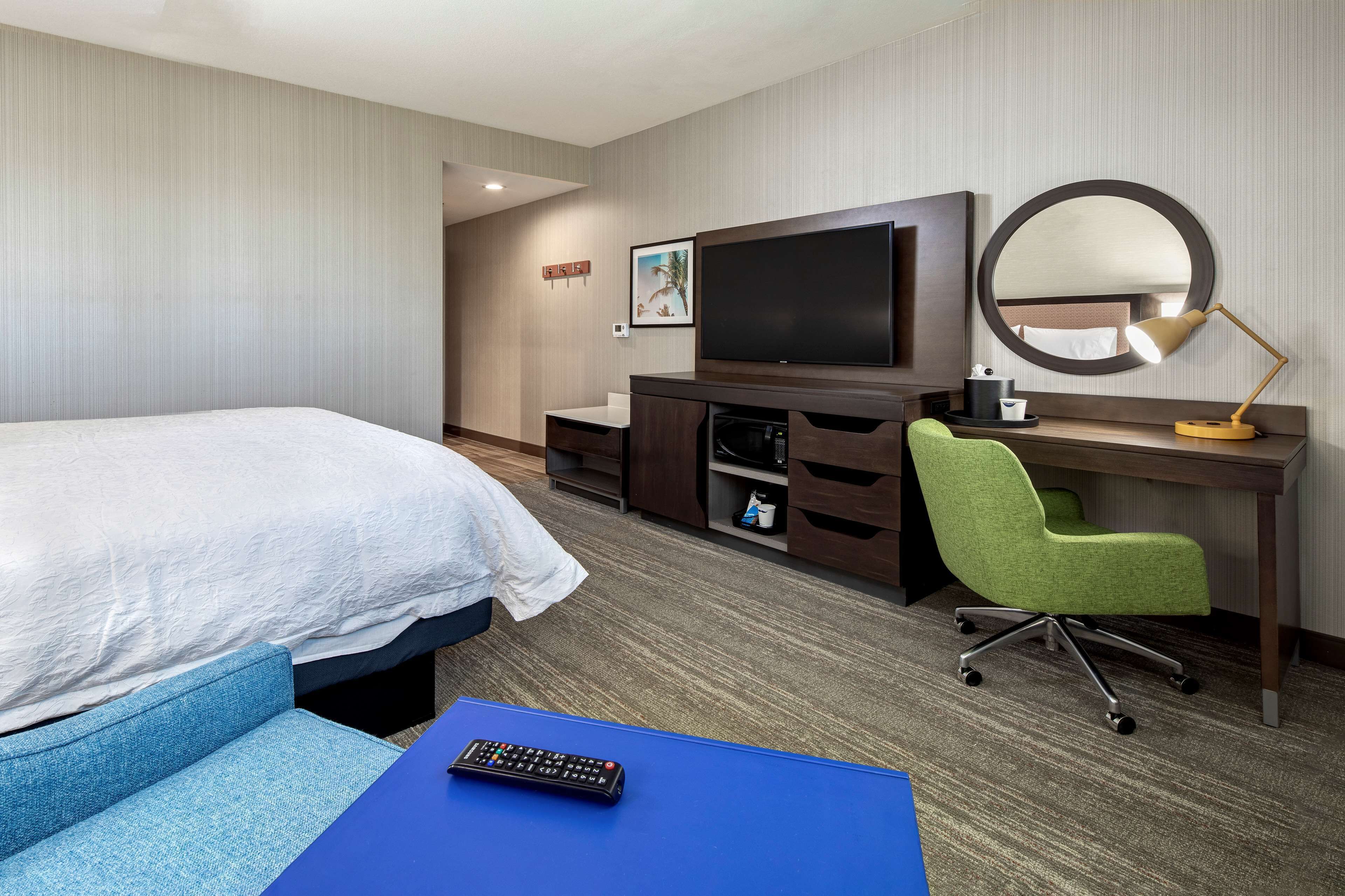 A hotel room with a bed, a desk with a computer, a chair, a TV, and a mirror.