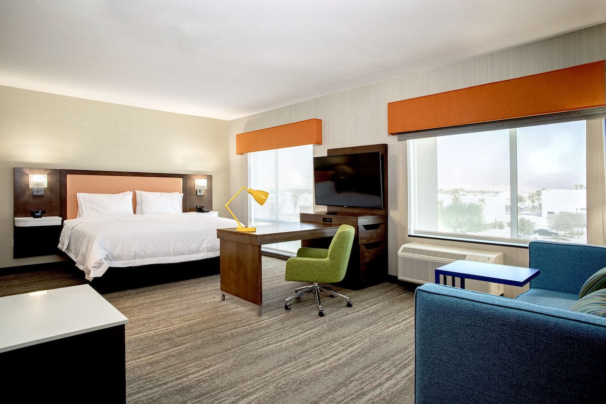 A hotel room with a bed, desk, chair, TV, and large windows offering a city view.