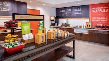 Zona de desayuno tipo buffet con una variedad de alimentos y bebidas, incluyendo un cartel de zumo recién exprimido y una presentación de gofres.