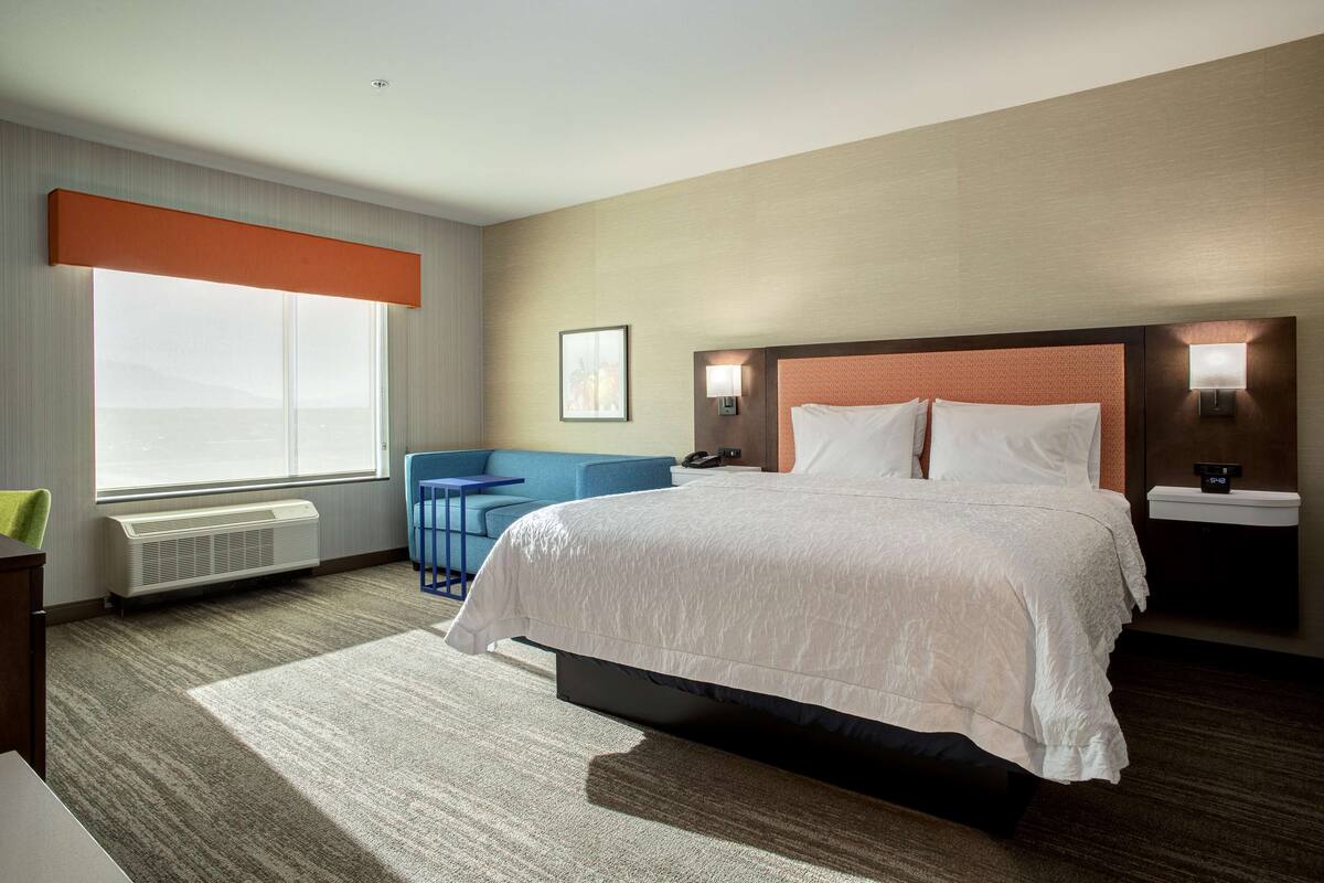 A hotel room with a large bed, a desk, a chair, a window, and a wall-mounted lamp.