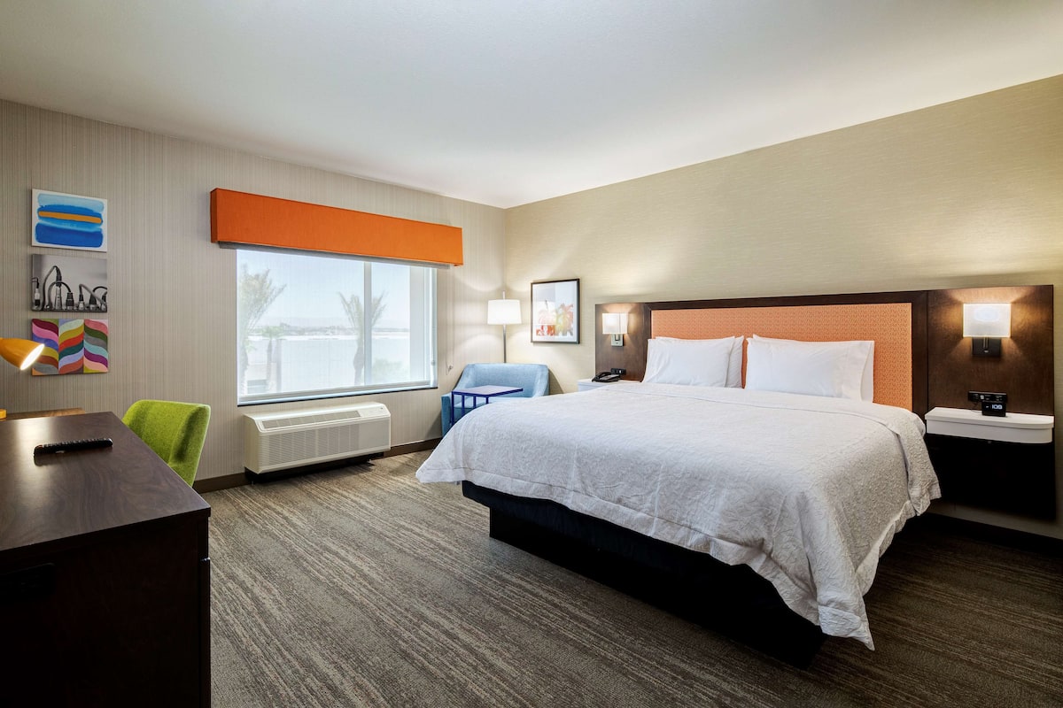 A hotel room with a large bed, a desk, a chair, a window with a view, and a wall with artwork.