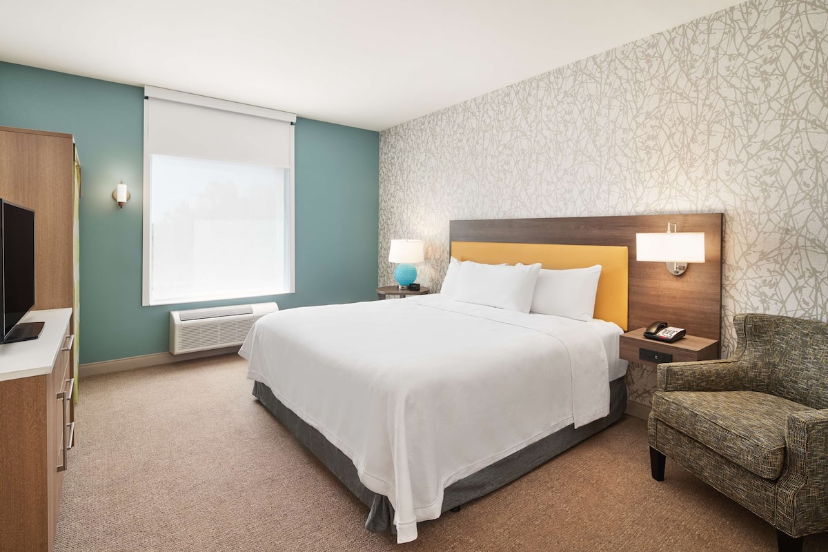 Une chambre d’hôtel avec un grand lit, un bureau doté d’un téléviseur à écran plat, une chaise et une fenêtre avec des stores.