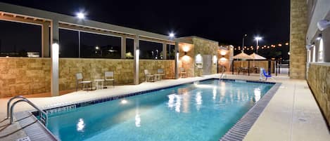 A swimming pool with a tiled edge, a safety ladder, and a lifeguard chair. There are lounge chairs and umbrellas around the pool area.