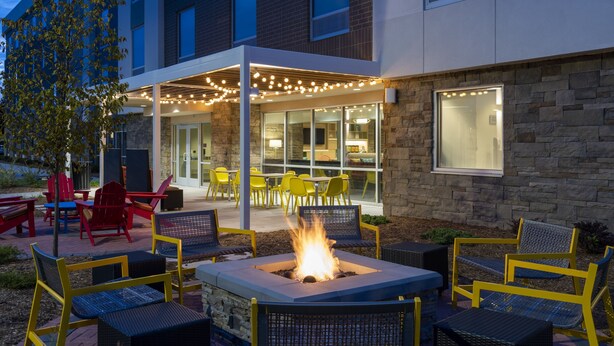An outdoor seating area with fire pit, yellow chairs, and tables, surrounded by a building with stone walls and large windows. - Home2 Suites by Hilton Palm Bay Melbourne I 95 (Palm Bay)