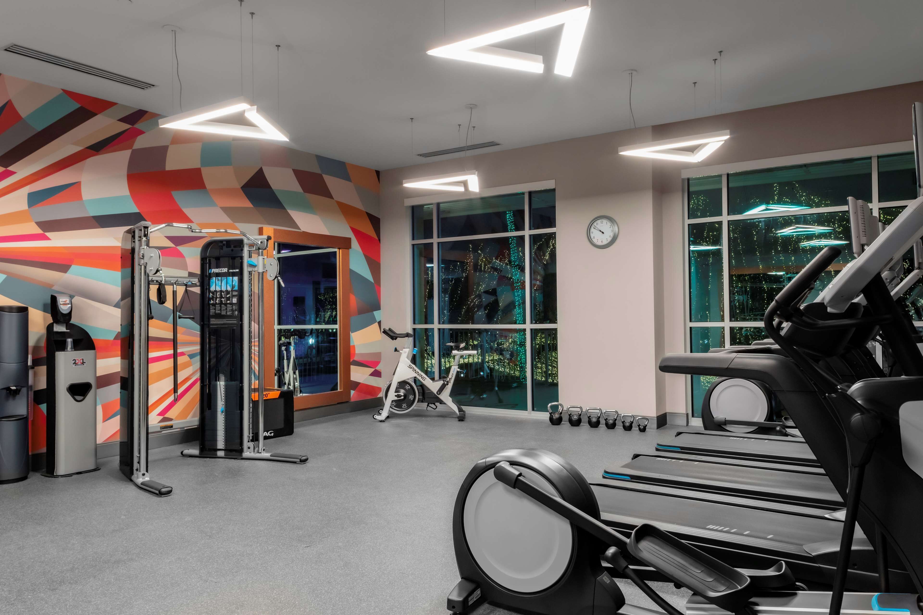 A gym with exercise equipment, a colorful wall, and large windows.