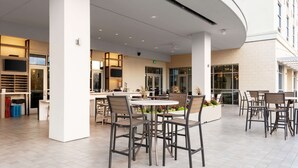 A modern outdoor dining area with round tables and chairs, surrounded by large windows and a white exterior wall. - Hilton Garden Inn Ocala Downtown (Ocala)