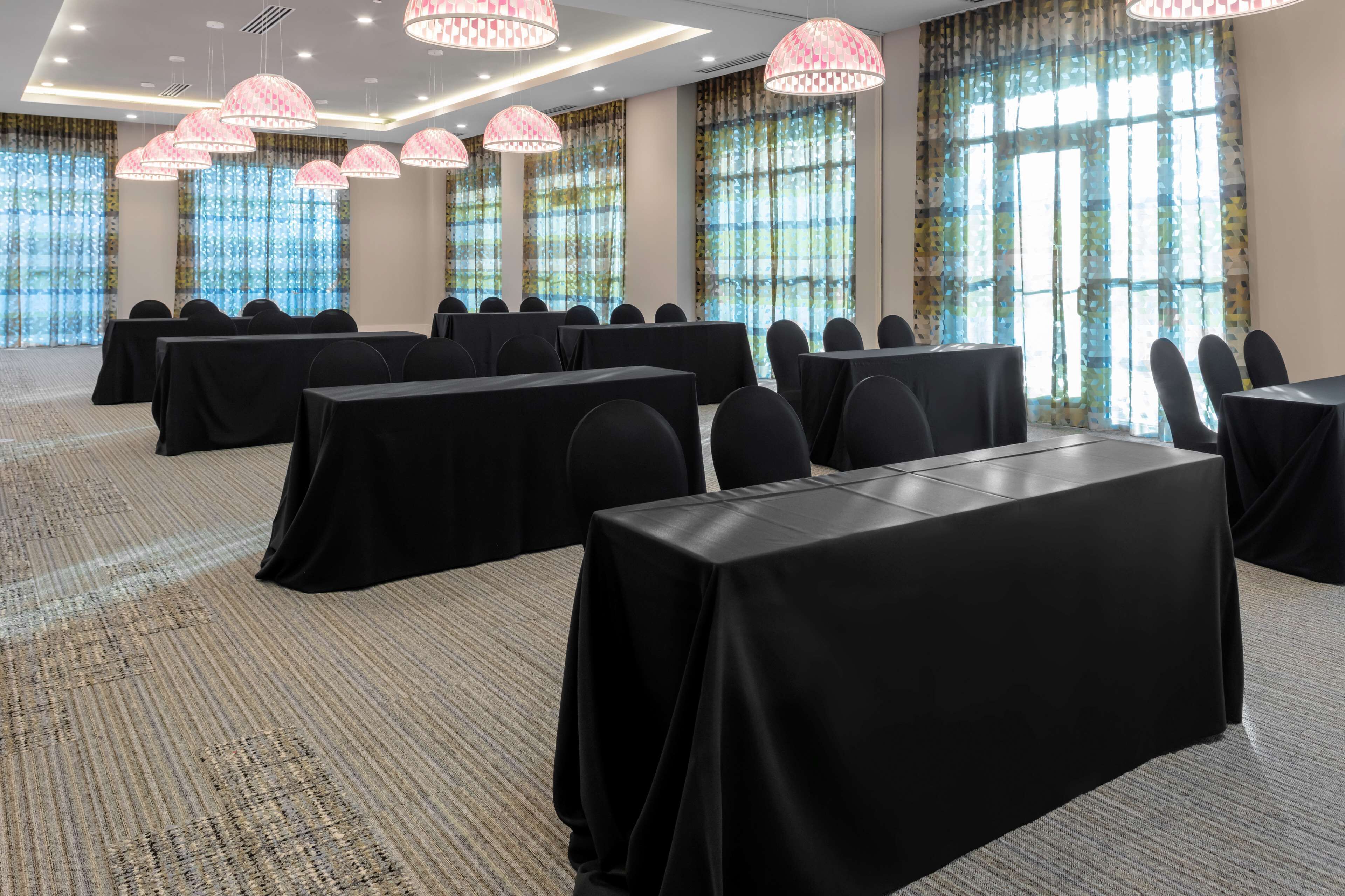 Un salón de conferencias con mesas y sillas, ventanas grandes con cortinas y lámparas colgantes.