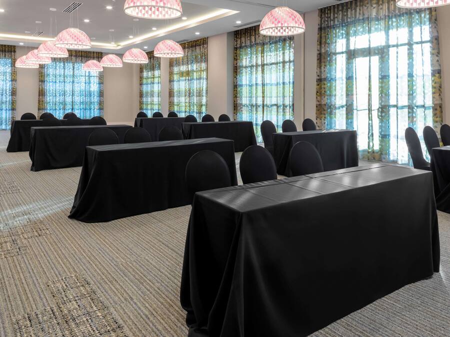 Un salón de conferencias con mesas y sillas, ventanas grandes con cortinas y lámparas colgantes.