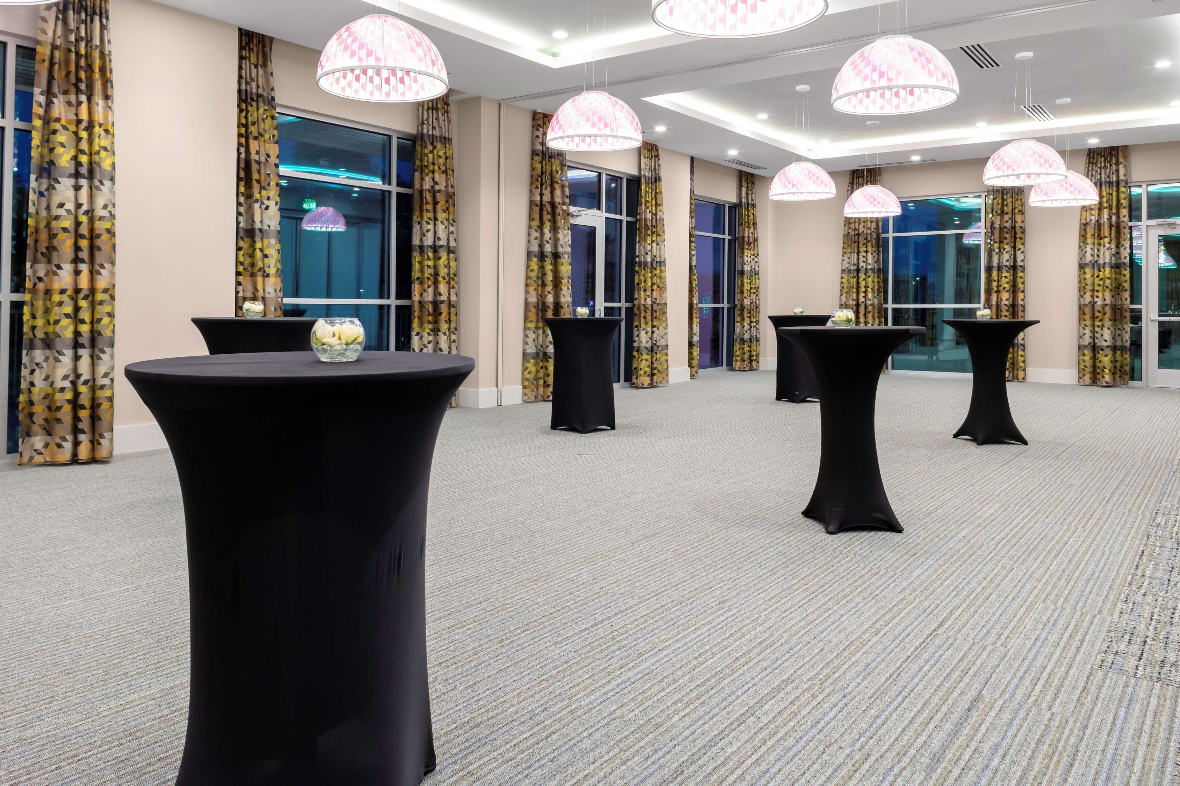 A spacious room with multiple round tables and black chairs, large windows with patterned curtains, and hanging pendant lights.