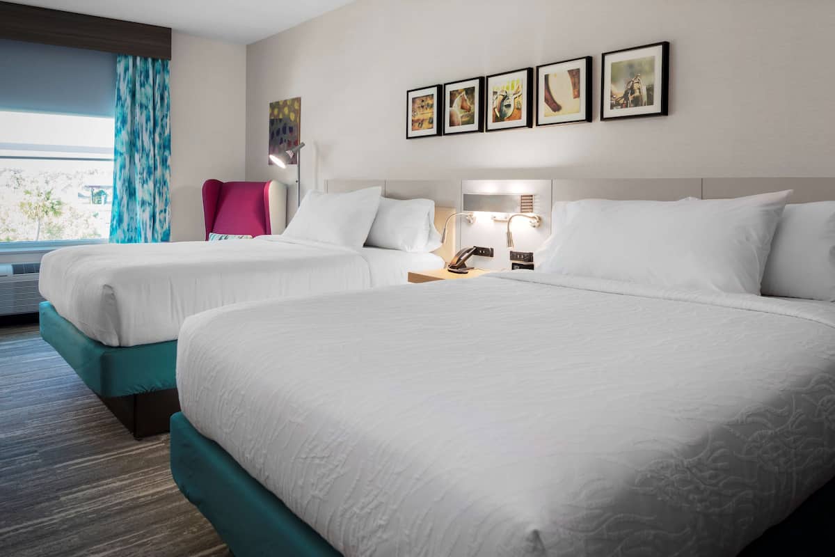 A hotel room with two beds, a desk with a lamp, and a window with curtains.
