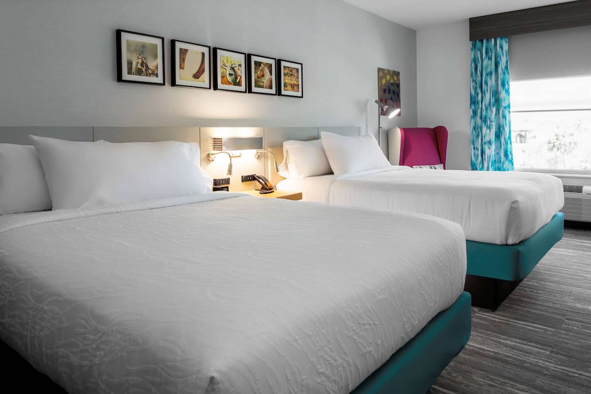 A hotel room with two beds, a nightstand, a lamp, and a window with curtains.
