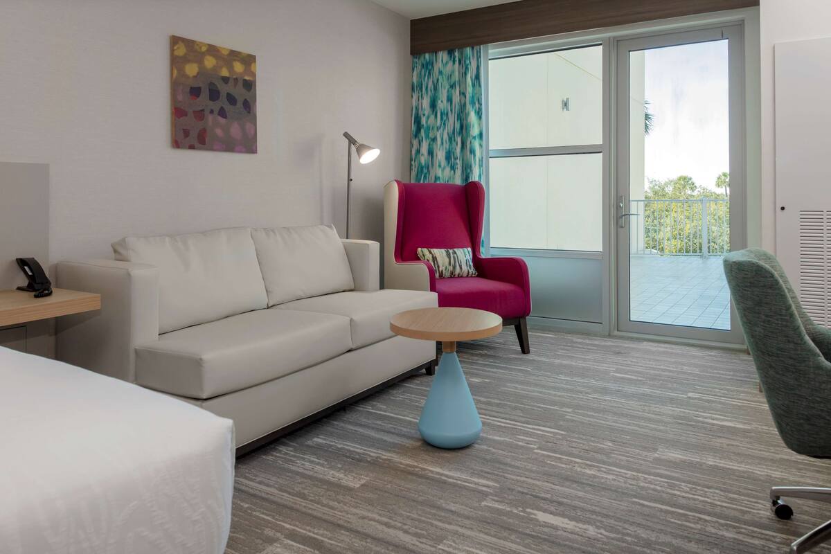 A modern hotel room with a sofa, a pink armchair, a round table, and a large window with a view of the outdoors.