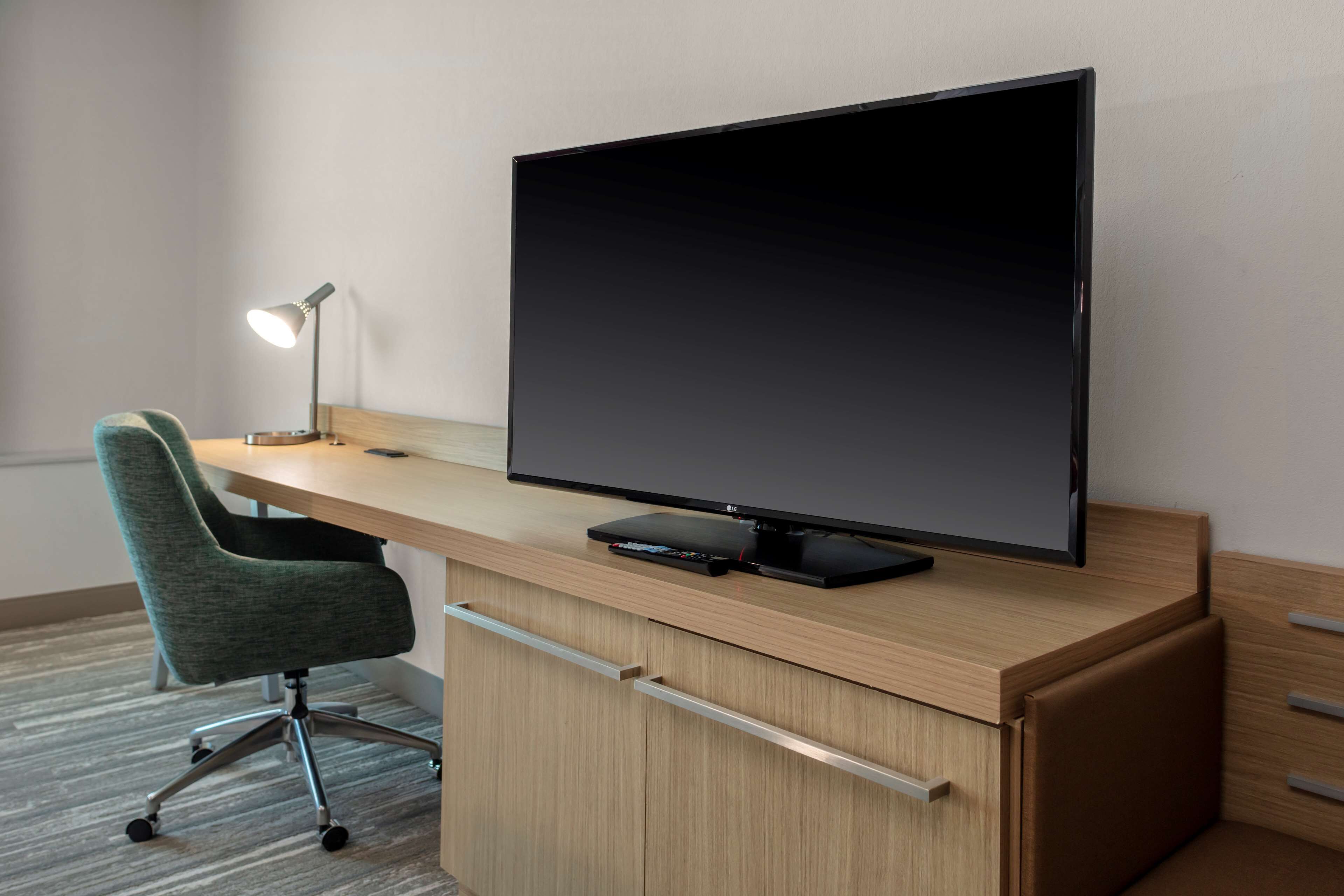 Une chambre d’hôtel moderne avec un bureau en bois, un téléviseur à écran plat, un fauteuil de bureau vert et une lampe de bureau.