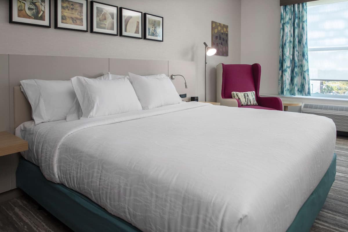 A neatly made bed with white linens, a headboard, and a teal accent at the foot. There is a red chair with a pillow, a bedside table, and a lamp.