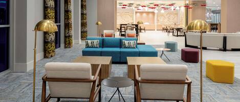 Een moderne lobby met een blauwe bank, beige fauteuils en een kleine ronde tafel. Er staan vloerlampen, een geel pouf en een muur met een patroon.