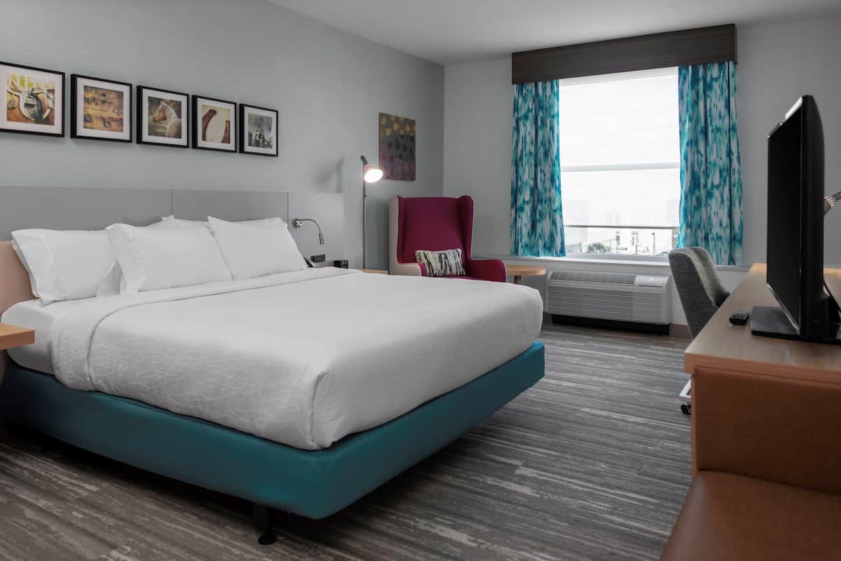A hotel room with a large bed, a desk with a TV, a chair, a desk lamp, and a window with curtains.