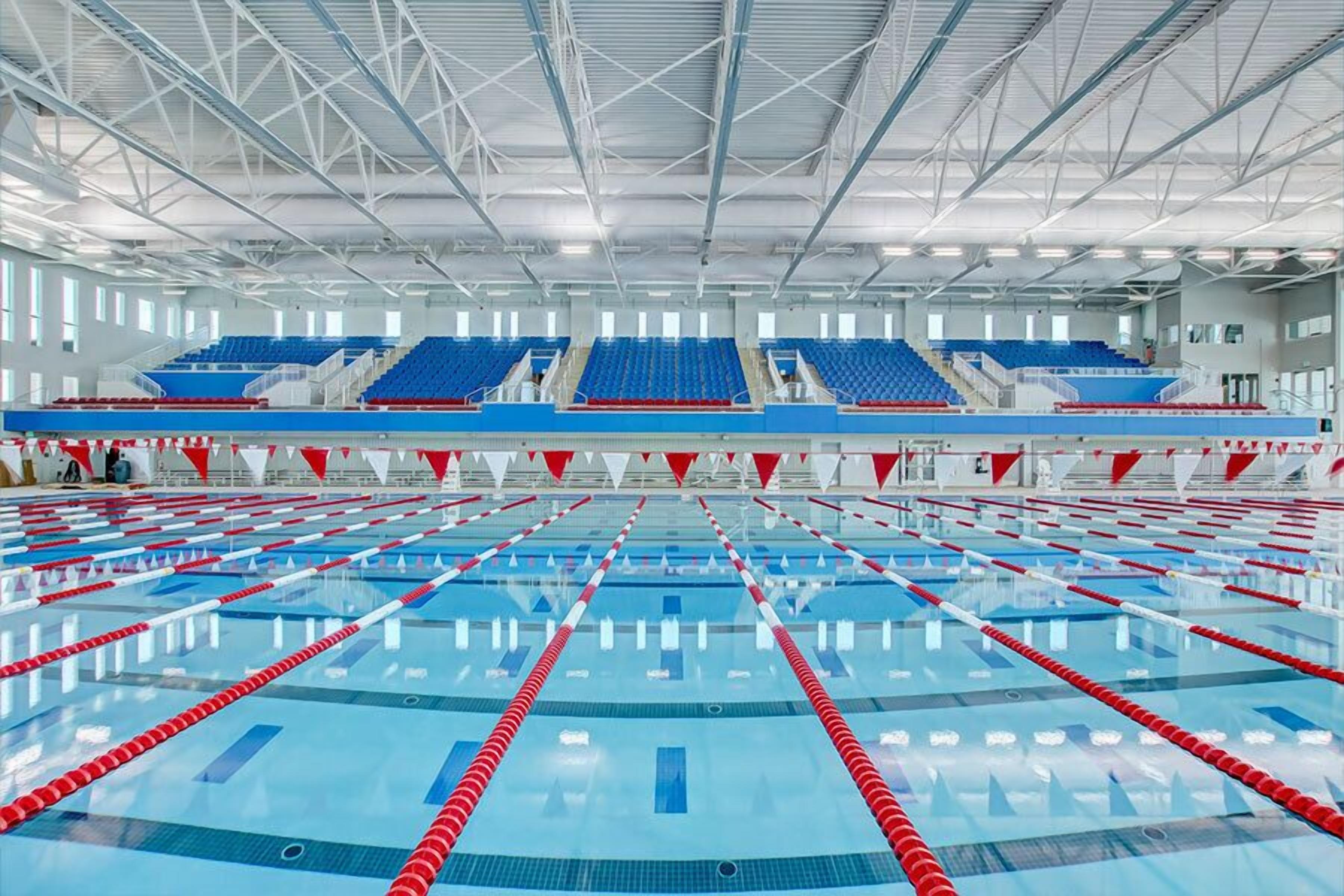 Une grande piscine intérieure dotée de plusieurs couloirs, de gradins pour les spectateurs et d’un plafond haut avec des poutres apparentes.