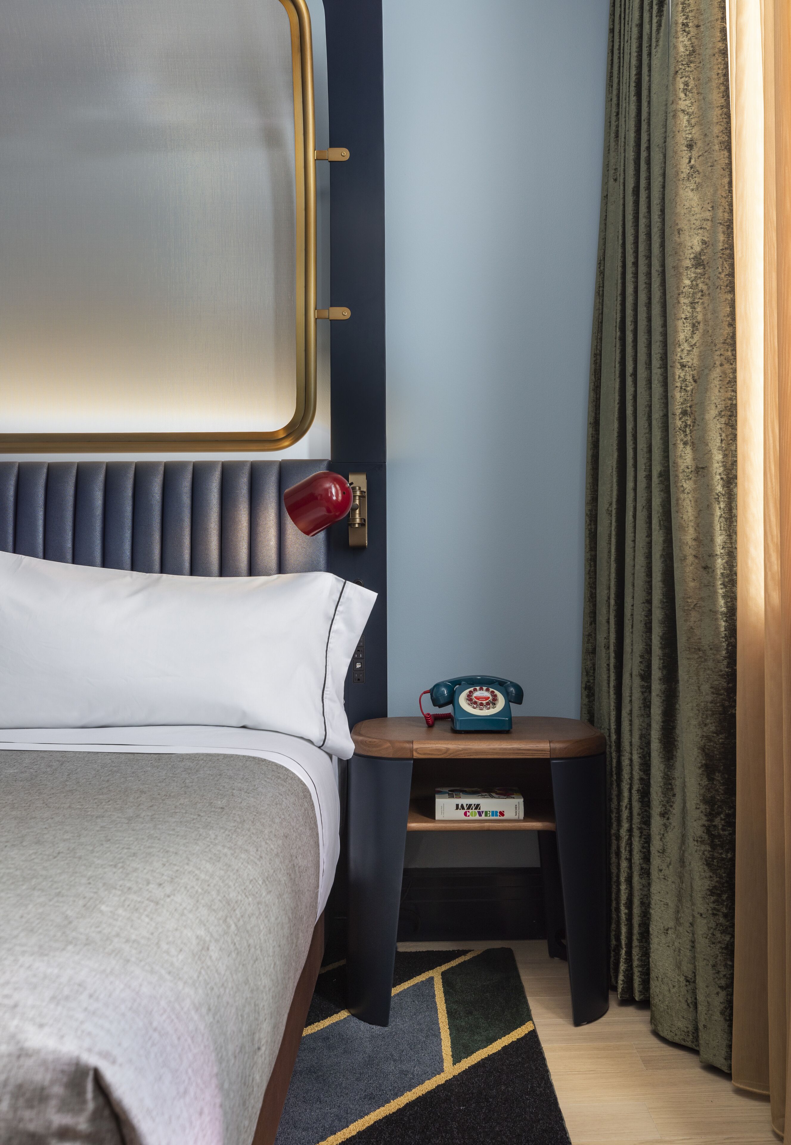 A neatly made bed with a grey blanket, a bedside table with a vintage-style telephone, and a small box labeled "KAZOO COVERS" on the table.