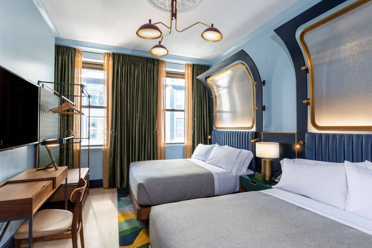 A hotel room with two beds, a desk, a chair, a TV, and a large window with curtains.