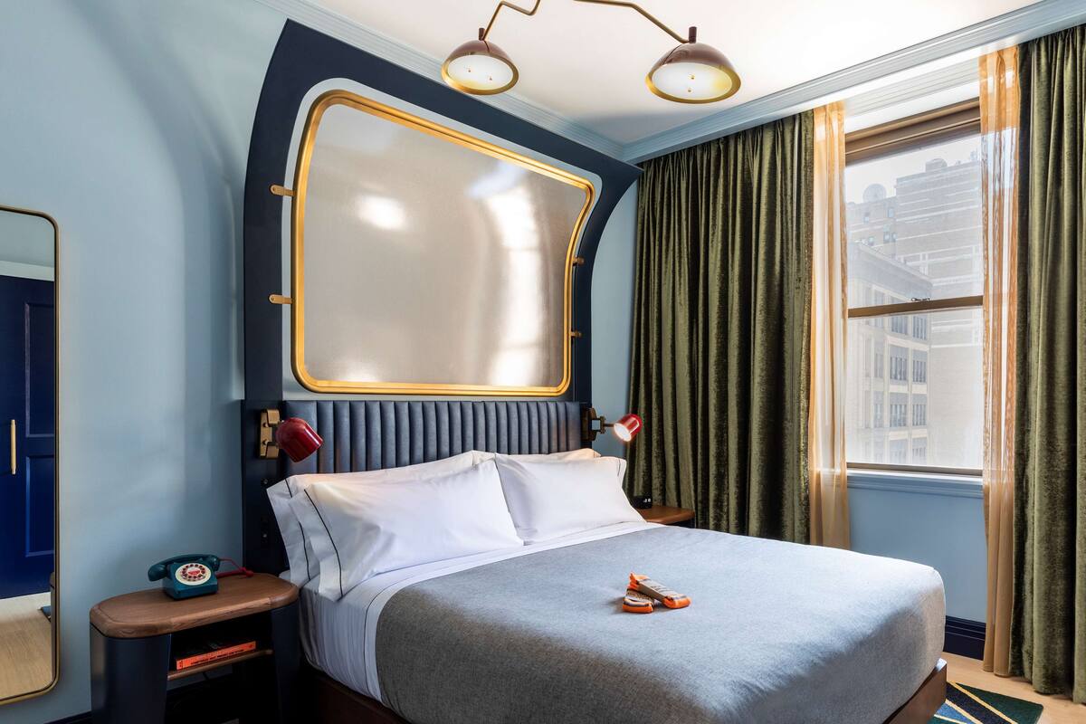 A hotel room with a large bed, a bedside table with a telephone, a mirror, and a window with curtains.