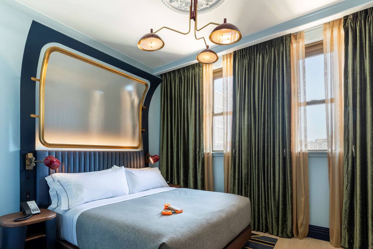 A hotel room with a large bed, a bedside table with a telephone, a decorative mirror, and a window with curtains.