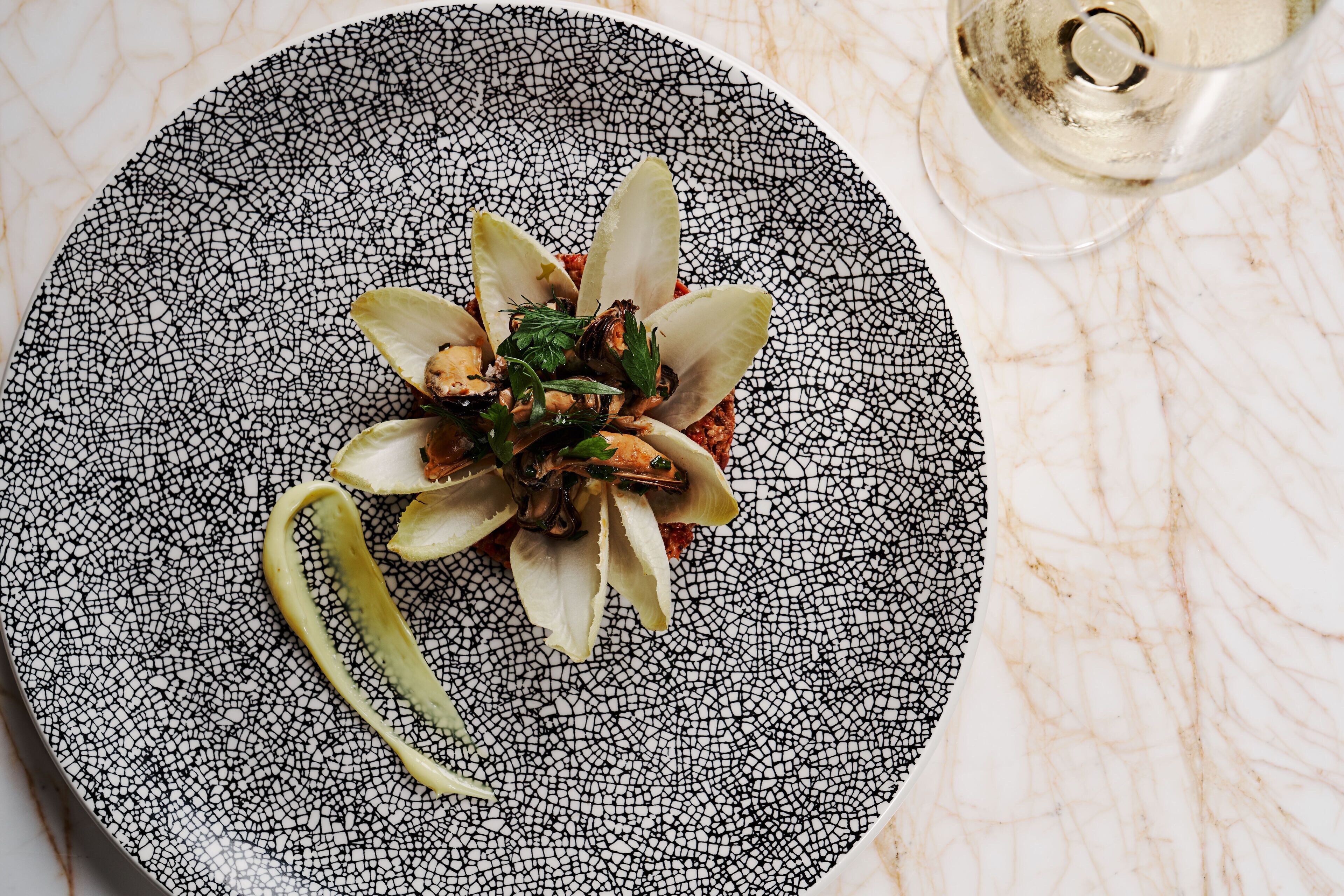 A plate with a dish on a textured black and white plate, a glass of white wine, and a marble surface.
