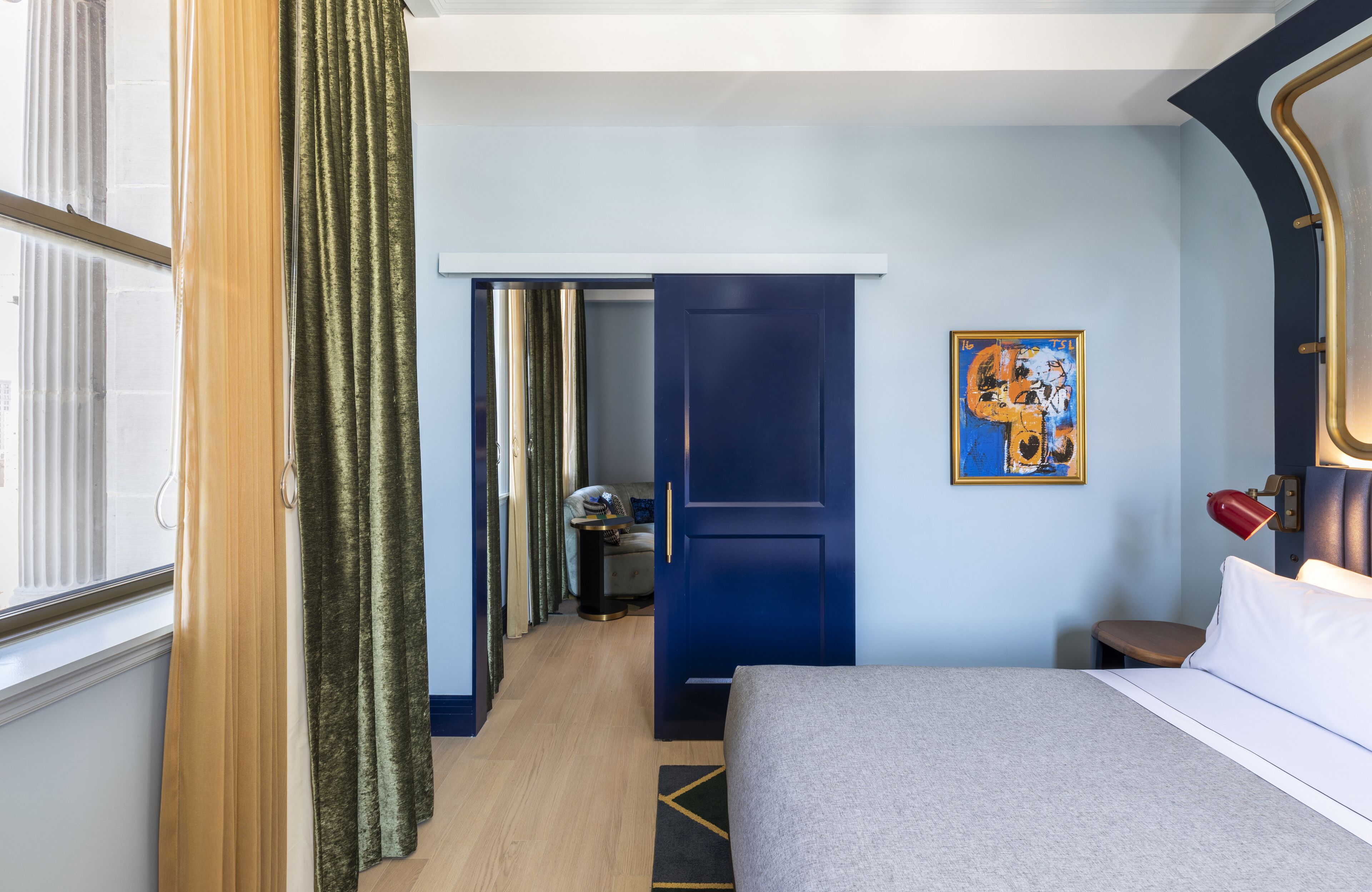 A hotel room with a bed, a desk, a chair, a painting on the wall, and a door leading to another room.
