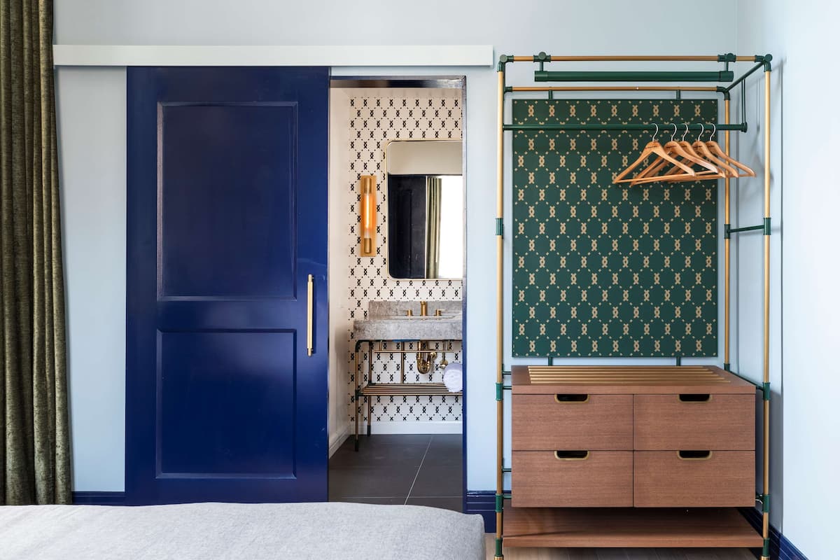 A hotel room with a blue door, a wooden dresser, and a hanging rack with clothes.