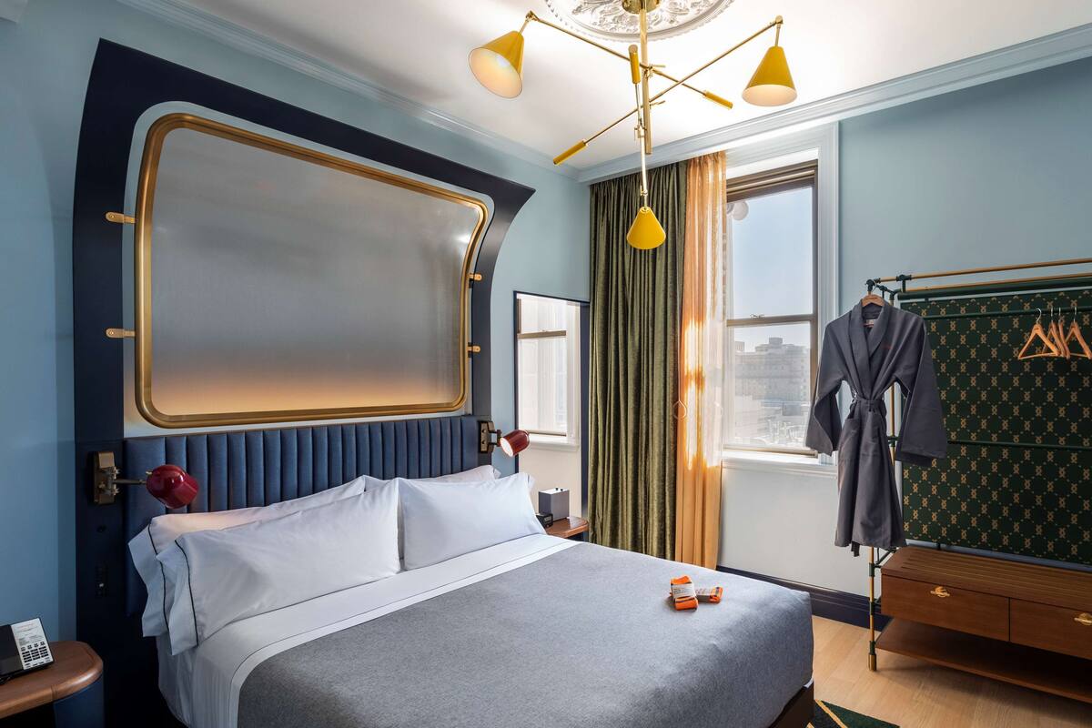 A hotel room with a large bed, a bedside table with a lamp, a window with curtains, a robe hanging on a rack, and a green patterned wall.