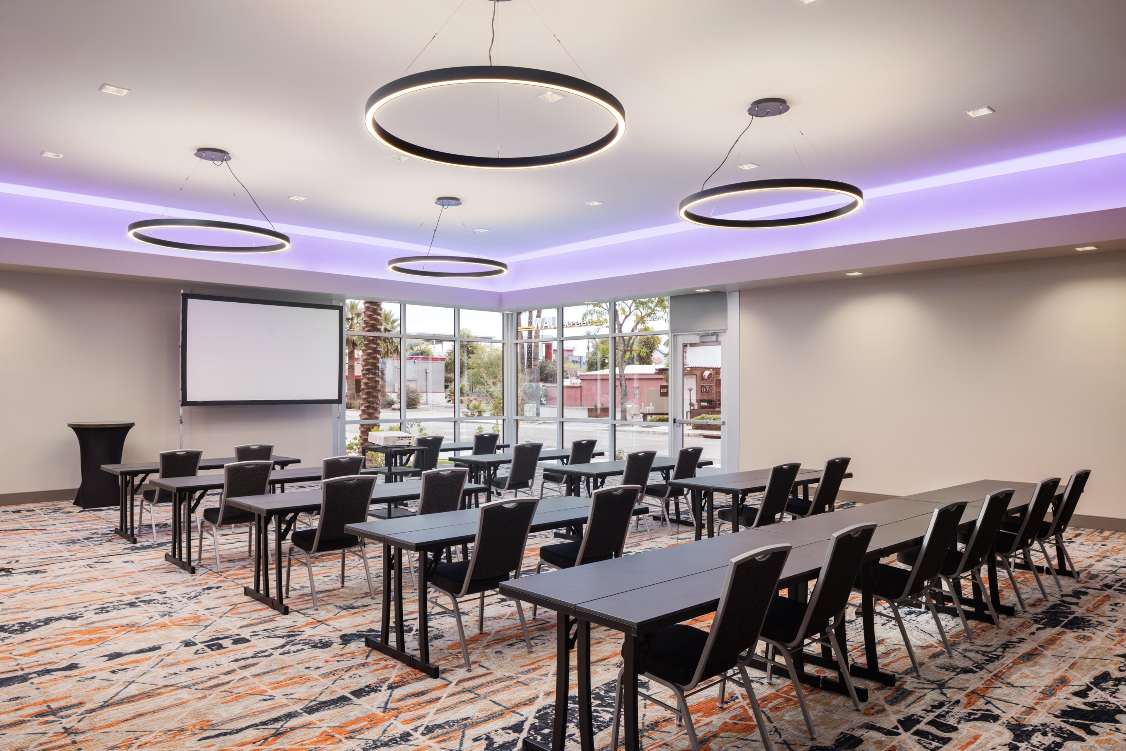 Une salle de conférence équipée de tables, de chaises, d’un écran de projection et de grandes fenêtres.