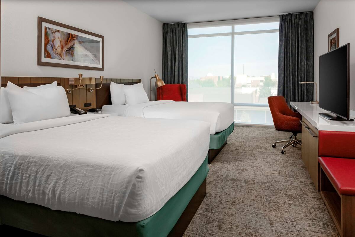 A hotel room with two beds, a desk, a TV, and a large window with curtains.