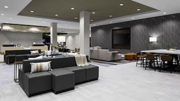 A modern hotel lobby with a sectional sofa, a dining area, and a kitchen in the background.