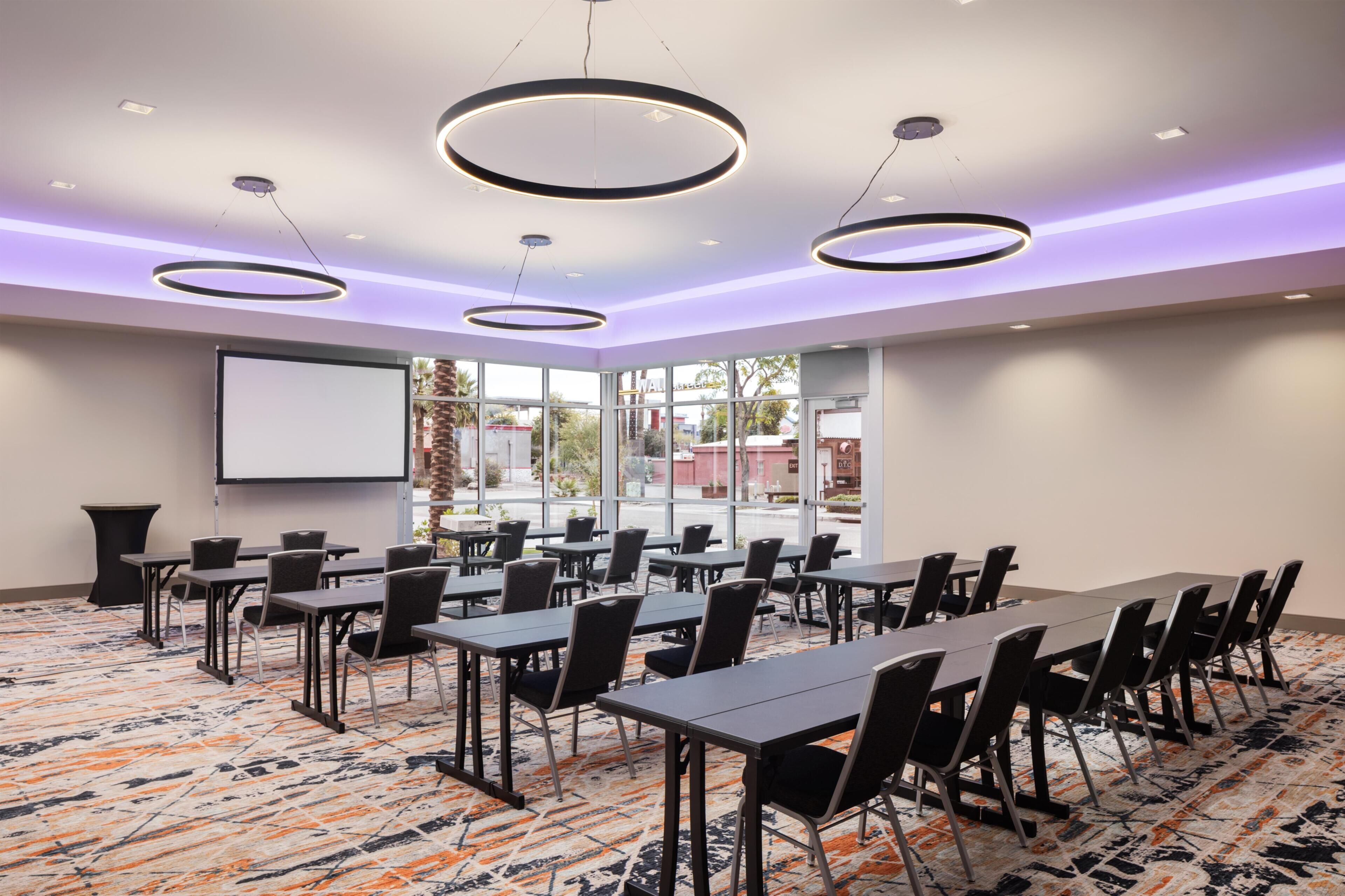 Un salón de conferencias con mesas, sillas, una pantalla para proyector y ventanas amplias.