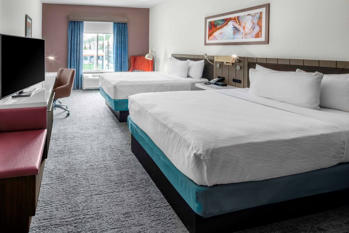 A hotel room with two beds, a desk with a TV, a chair, and a window with curtains.
