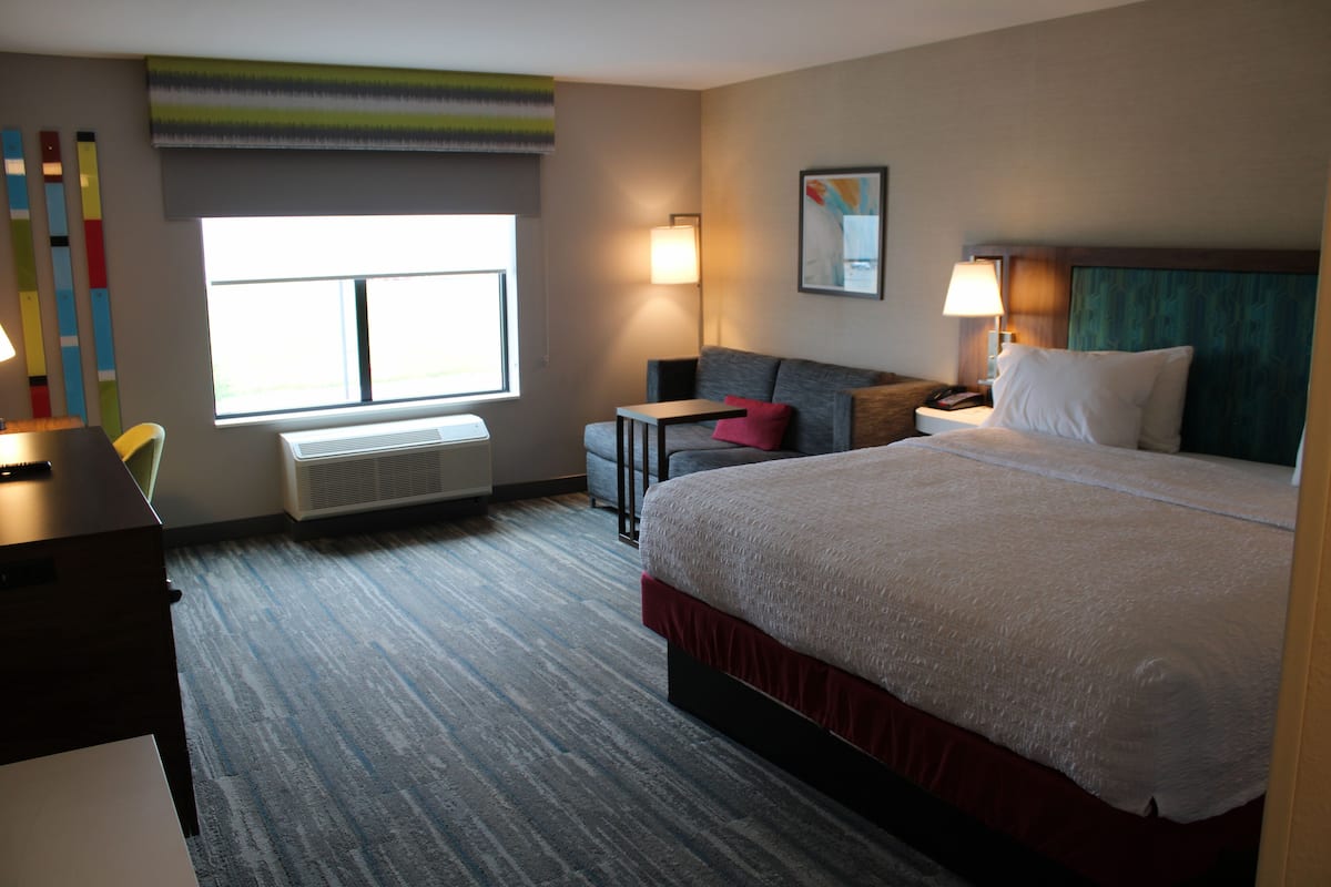 A hotel room with a large bed, a desk, a chair, a small table, and a window with blinds.