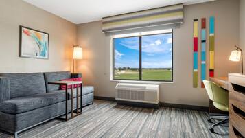 A hotel room with a grey sofa, a small table, a window with a view of a green field, a radiator, and a colorful wall decoration.